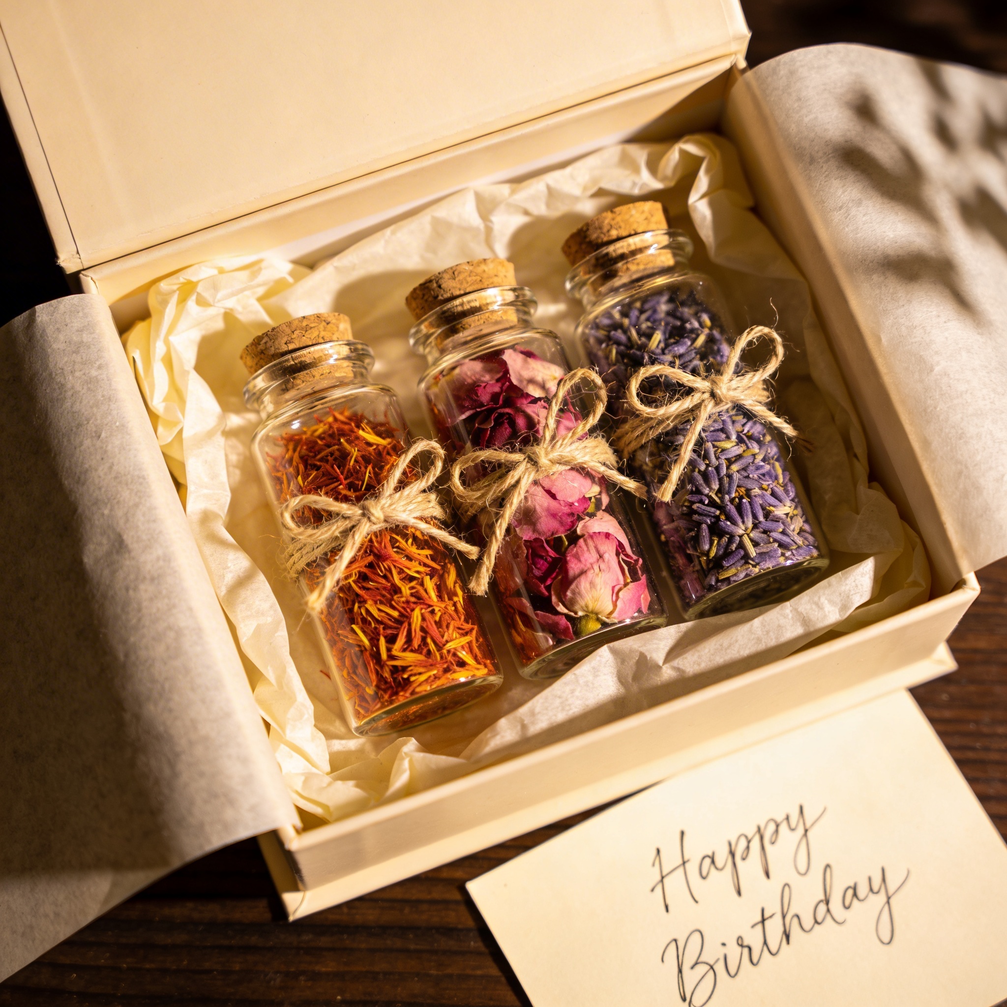 Gift box with cream tissue paper containing three glass jars of saffron, rose, and lavender, linen ribbon